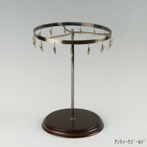 ●商品名:チーフ・ハンカチスタンド回転式 木製ベース仕様 ●表面処理:アンティークゴールドメッキ(AG)仕上 ●寸法:高さ315mm 上部回転リング直径240mm ●上部回転式リング:クリップ15個付 ●材質:スチール ●特長:百貨店様では棚板高さのスパンが30cm強の設定が多いと教えてもらい、では「高さ30cmのスペースにフィットするチーフツリーを作ろう」ということで高さ30cmのチーフツリーを製造しました。また、クリップの先端には塩ビのキャップを装着し、展示商品を傷つけにくい仕上がりになっています。さらに、木製のベースを組み合わせることで、落ち着きのある存在感と高級感を演出できます。 ●生産国:日本(タヤ自社工場)