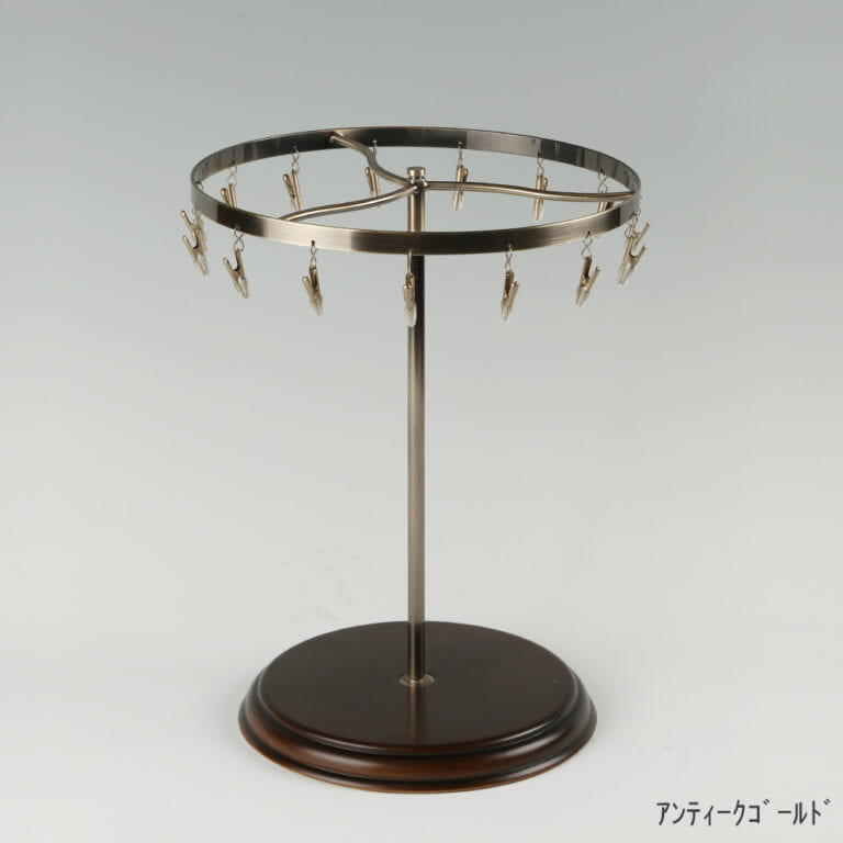 ●商品名:チーフ・ハンカチスタンド回転式 木製ベース仕様
●表面処理:アンティークゴールドメッキ(AG)仕上
●寸法:高さ315mm 上部回転リング直径240mm
●上部回転式リング:クリップ15個付
●材質:スチール
●特長:百貨店様では棚板高さのスパンが30cm強の設定が多いと教えてもらい、では「高さ30cmのスペースにフィットするチーフツリーを作ろう」ということで高さ30cmのチーフツリーを製造しました。また、クリップの先端には塩ビのキャップを装着し、展示商品を傷つけにくい仕上がりになっています。さらに、木製のベースを組み合わせることで、落ち着きのある存在感と高級感を演出できます。
●生産国:日本(タヤ自社工場)